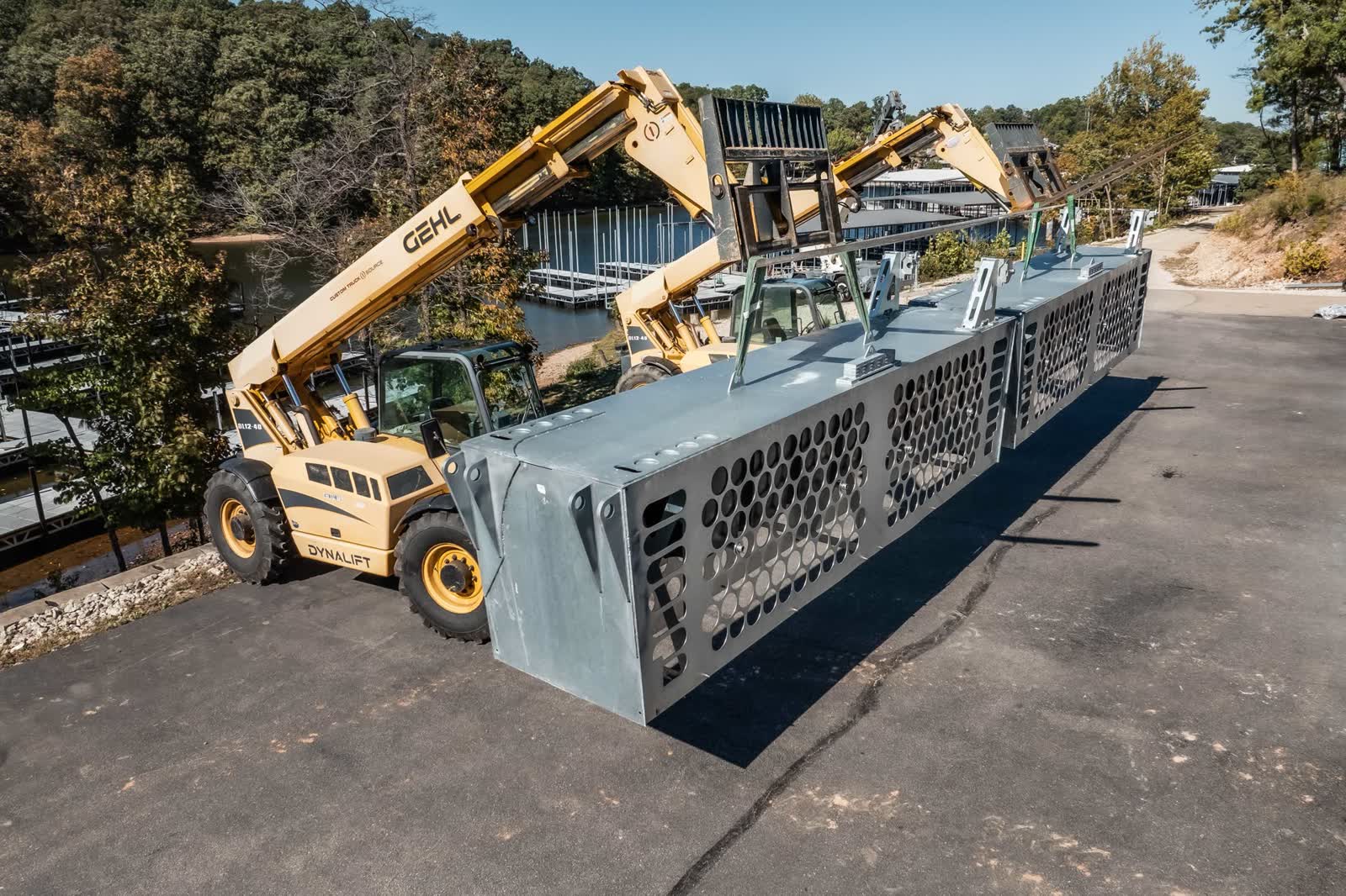 Telehandlers lifting a pair of galvanized WaveBaffle sections on a lakeside lot, with covered marina docks visible behind the trees